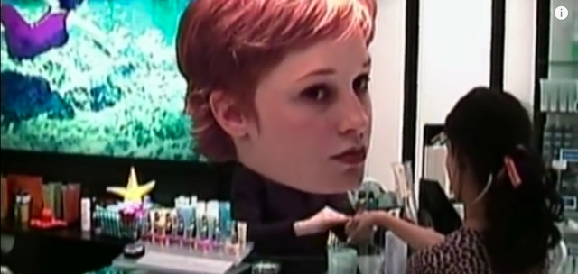 Screenshot of the heroine attending to customers at her cosmetics counter, complete with her own oversized cut-out of her face from the video "Let Forever Be."